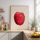 Framed art print of a raspberry on a kitchen wall with a window and shelves in the background.