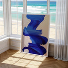 Abstract blue art print on a stand in a room with large windows showing a beach view.