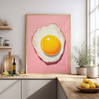 Framed art print of a fried egg on a pink background in a kitchen setting.
