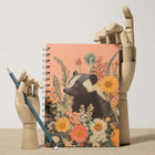 Notebook with a badger illustration surrounded by flowers, held by a wooden hand with a pencil.
